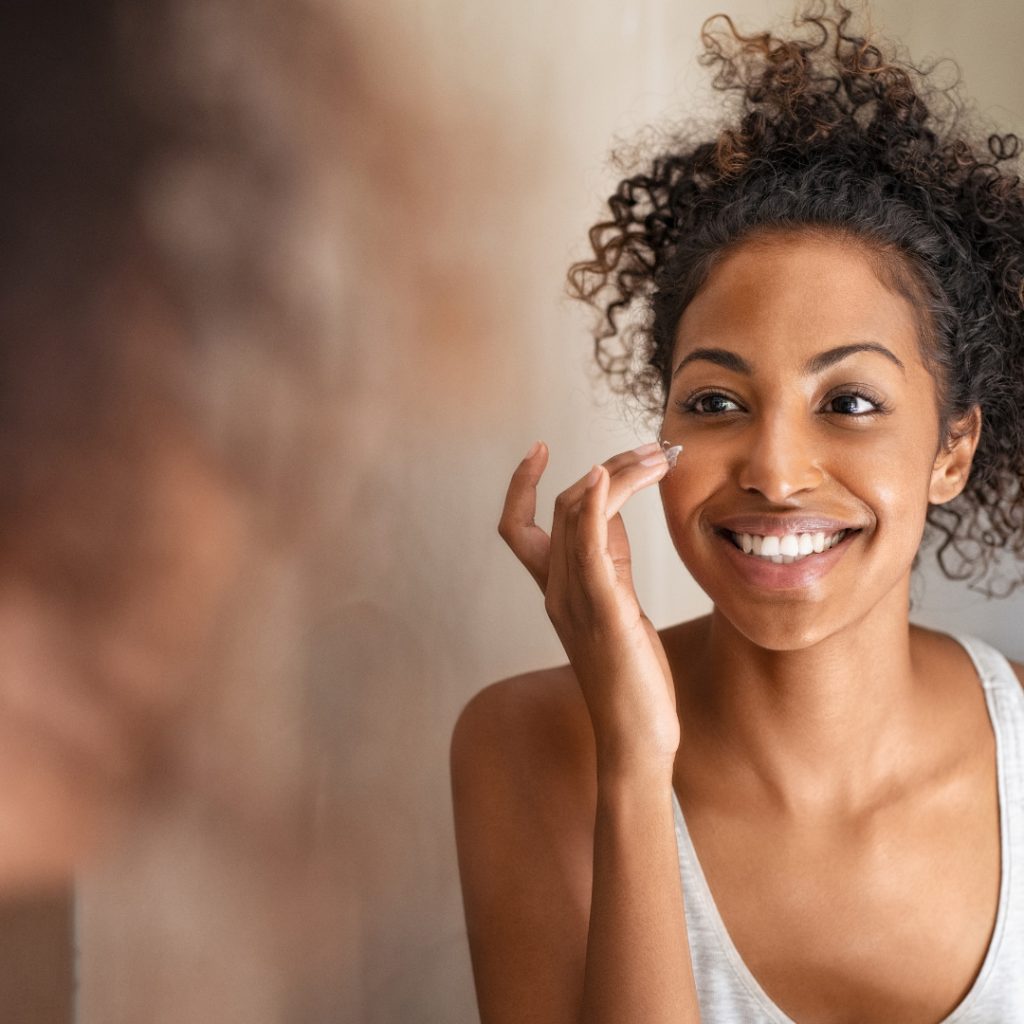 Black woman putting face cream on check and smiling in mirror